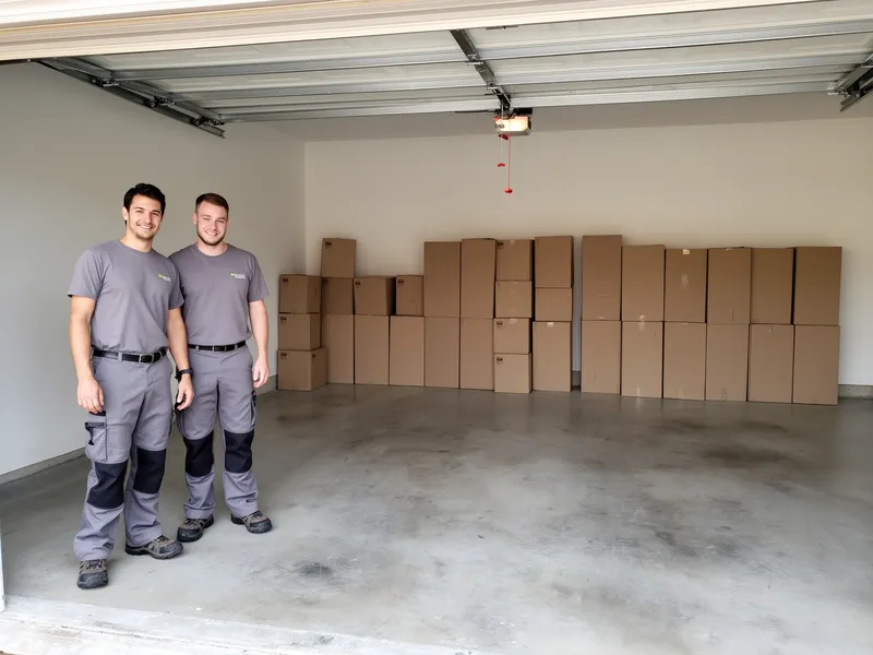 Zwei Fachkräfte in grauer Arbeitskleidung stehen in einer leeren, sauberen Garage mit ordentlich gestapelten Kartons.